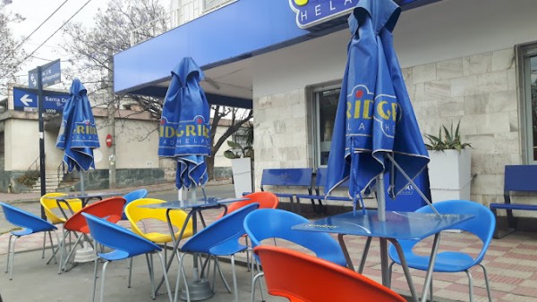 Mesas y sillas coloridas bajo sombrillas azules en terraza de café. Ambiente urbano con árboles deshojados al fondo.