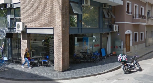 Pizzería Moma: fachada moderna con mesas afuera, gente caminando y moto estacionada. Ambiente urbano soleado.