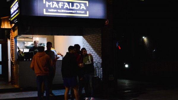 Noche en Mafalda: gente espera entrada, iluminación cálida, ambiente animado, fachada de ladrillos.
