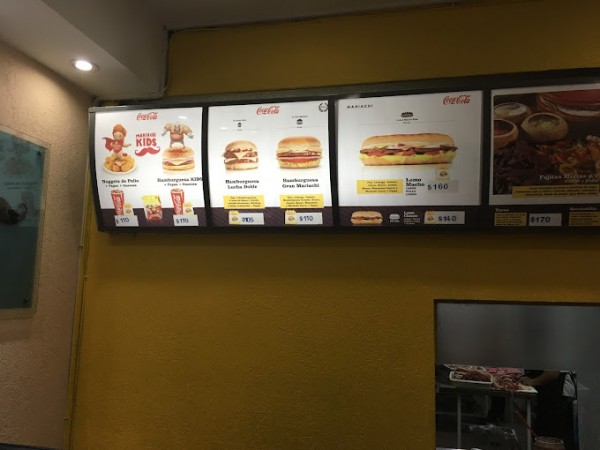 Cartel amarillo con menú de hamburguesas y bebidas en el restaurante Mariachi. Precios visibles. Decoración sencilla.