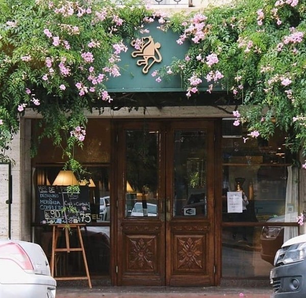 Entrada del Yolanda Hotel con puertas de madera tallada y fachada cubierta de flores rosadas. Atmosfera acogedora.