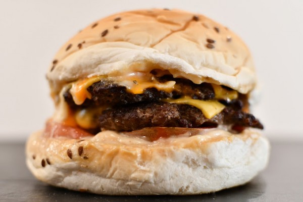 Una hamburguesa doble con panecillo de semillas de sésamo. La hamburguesa tiene dos jugosas carnes de res, cada una cubierta con queso derretido. Entre las carnes se puede ver una rebanada de tomate y salsa de tomate. La hamburguesa está montada en un pan