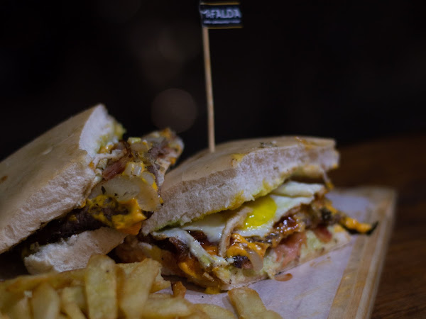 Sandwich de Mafalda con huevo, queso y cebolla, acompañado de papas fritas. Bandera negra con nombre del lugar.