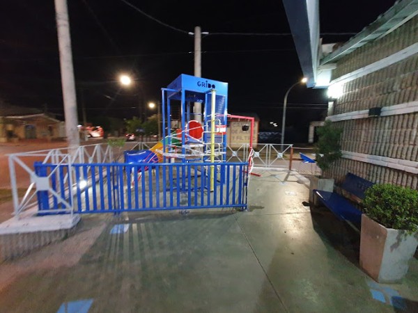 Parque infantil nocturno con estructura azul, tobogán y barrera metálica. Farolas iluminan el área.