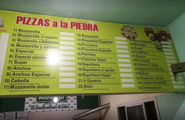Cartel amarillo con lista de pizzas a la piedra en Montagú. Variedad de sabores y precios. Decoración sencilla.