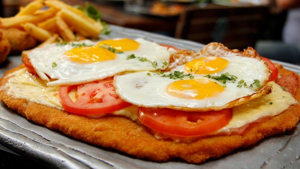 Milanesa con huevos fritos y tomate, servida en bandeja metálica. 