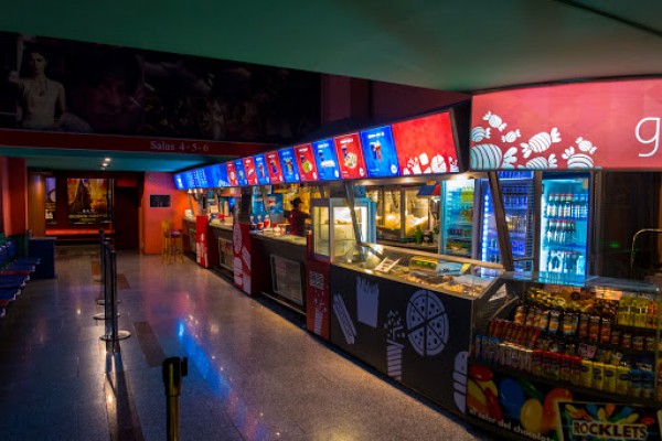Interior del Gran Rex: pasillo con taquillas iluminadas, máquinas expendedoras y carteles coloridos. Ambiente vibrante y acogedor.