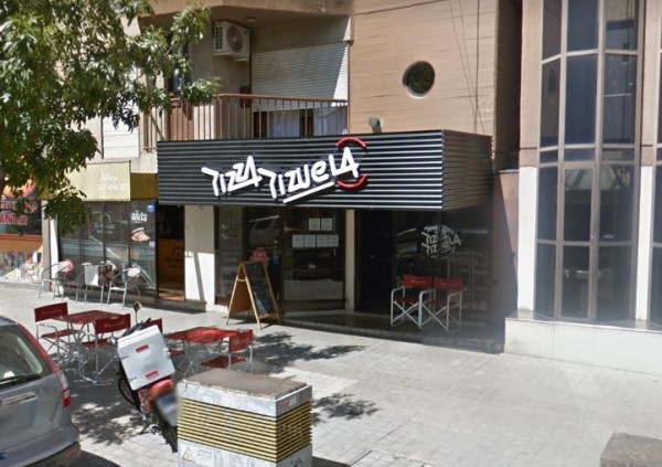 Fachada de Pizza Pizuela con sillas rojas afuera, cartel negro y blanco, árbol y bicicleta cerca.