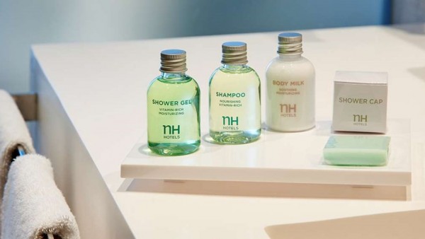 NH Urbano: Toallas blancas y productos de baño verdes y blancos sobre superficie blanca. Gel, champú, leche corporal y gorro de ducha.