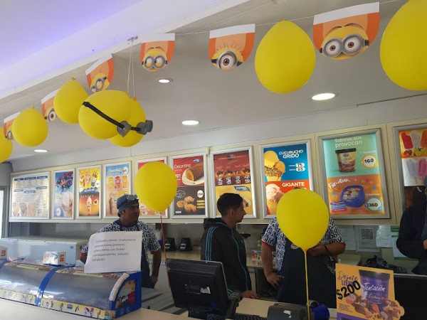 Interior de heladería con globos amarillos y carteles de Minions colgando del techo. Clientes y empleados interactúan.