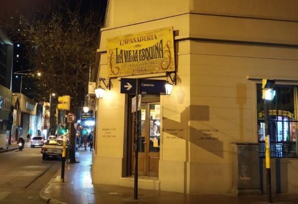 Noche en La Vieja Esquina: empanadería iluminada, calle animada, coches y peatones transitando. Estilo urbano clásico.