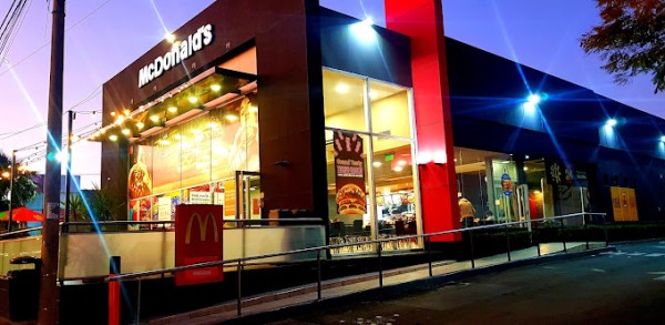 Restaurante McDonald's iluminado por la noche, con ventanas grandes y gente adentro. Decoración festiva en el exterior.