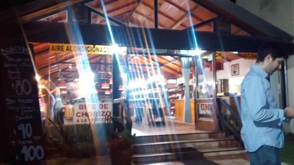 Entrada iluminada del Carmelo Parrilla Restaurante con gente adentro y carteles visibles. Ambiente acogedor y cálido.