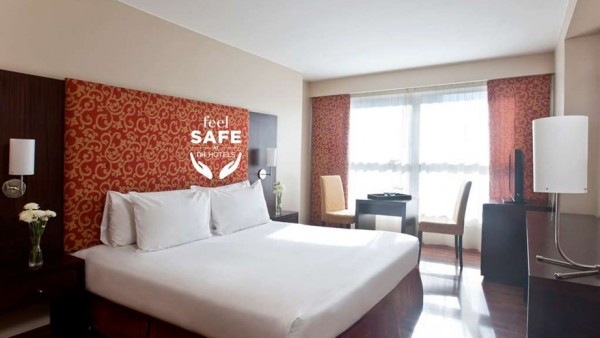 Habitación moderna de NH Urbano con cama grande, cortinas rojas y ventana panorámica. Ambiente acogedor y seguro.