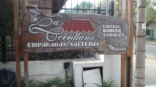 Cartel de madera con logo y texto anunciando empanadas salteñas, locro, humita y tamales. Fondo urbano.