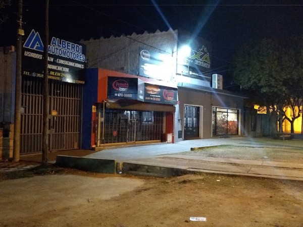Edificio nocturno con locales comerciales iluminados, incluyendo una empresa de automotores y un restaurante. Árboles y calle desierta.