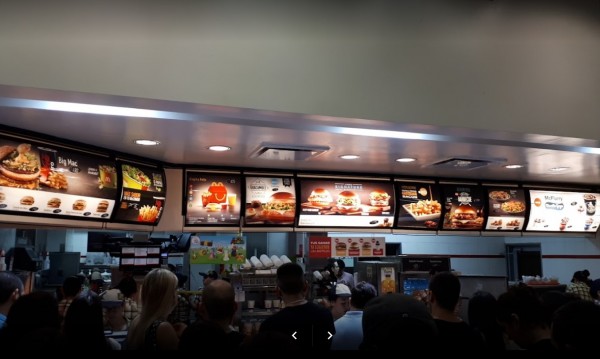 Interior de McDonald's con clientes esperando y menú iluminado en la pared. Ambiente ocupado y moderno.