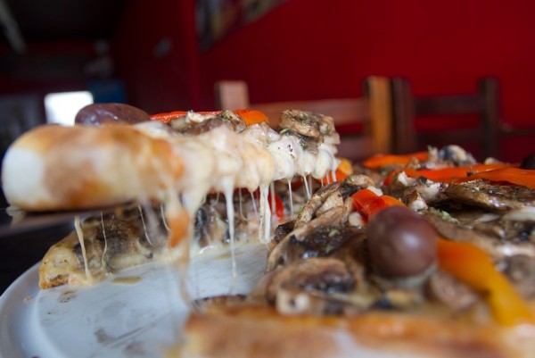 Pizza de Renato Pizza con champiñones, aceitunas y pimientos, rebanada con queso derretido. Fondo rojo.