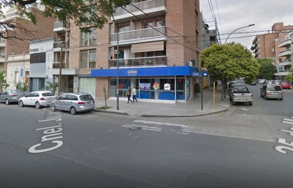 Edificio con fachada azul y blanca en esquina de calle. Personas caminando y autos estacionados frente a 