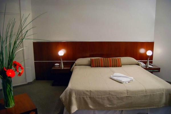 Habitación de 'El Virrey': cama con manta beige, almohada rayada, mesas laterales con lámparas y florero con flores rojas.