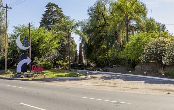Entrada al Hotel Ruta con camino pavimentado, vegetación exuberante y escultura con luna y letras rojas.