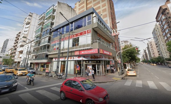 McDonald's en esquina urbana con edificios altos, coches y peatones transitando por la calle.