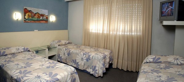 Habitación del Hotel Montecarlo con tres camas, cortinas beige y cuadro colorido en la pared.
