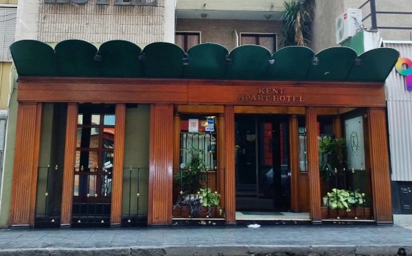 Entrada del Kent Apart Hotel con fachada marrón, techo verde y plantas decorativas. Edificio urbano con aire clásico.