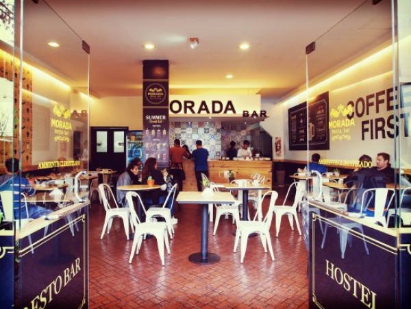 Interior acogedor de Morada Hostel con mesas blancas, sillas y gente disfrutando café. Ambiente cálido y acogedor.