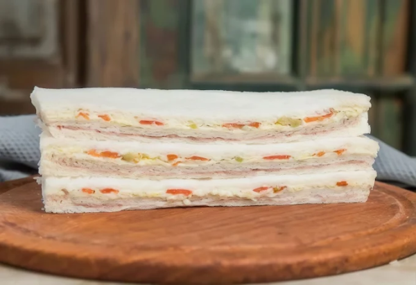 Tres sándwiches de miga triangulares apilados sobre tabla de madera circular, con relleno cremoso visible que incluye trozos de zanahoria naranja y verduras. Fondo con ventanas de madera.