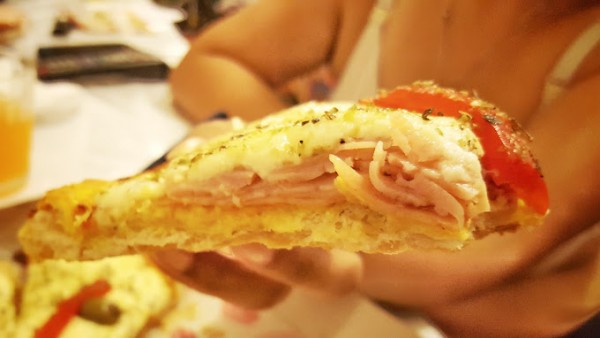 Rebanada de pizza con jamón y tomate en mano. Ambiente cálido de Comidas Homero.
