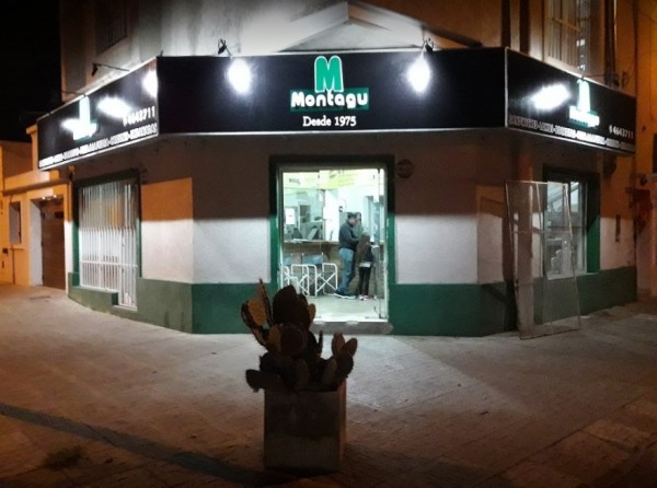 Tienda Montagú iluminada por la noche, fachada verde y blanca, letrero luminoso, personas dentro, ambiente urbano.