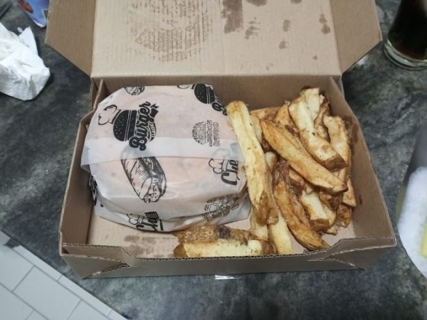 Una caja de cartón abierta contiene una porción de papas fritas doradas y crujientes junto a un envoltorio de papel blanco con ilustraciones de hamburguesas y texto. La caja está sobre una superficie de granito oscuro, y en el fondo se pueden ver parcialm