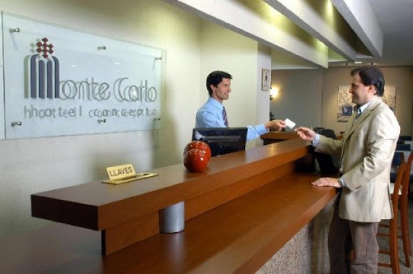 Recepción Hotel Montecarlo: empleado entrega llave a huésped en mostrador de madera. Ambiente moderno y acogedor.