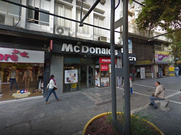 McDonald's en calle concurrida. Fachada blanca y roja, gente caminando. Árboles y tiendas alrededor. Atmosfera urbana.