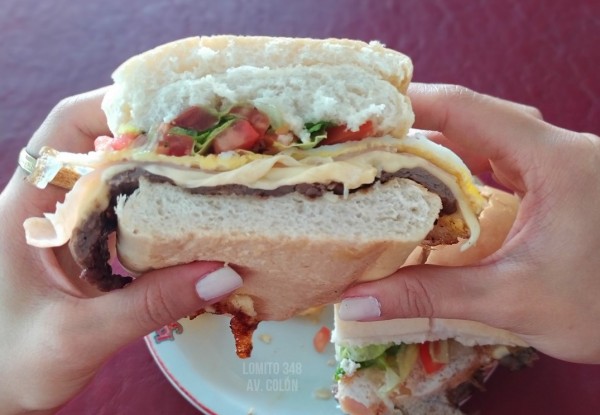 Manos sosteniendo hamburguesa con queso, tomate y lechuga en Lomitos 348. Fondo rojo.