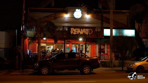 Noche en Renato Pizza: fachada iluminada, coches estacionados, gente dentro. Ambiente cálido y acogedor.