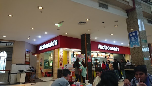 McDonald's en centro comercial: gente comiendo y caminando, mostradores con menú y empleados atendiendo. Iluminación brillante.