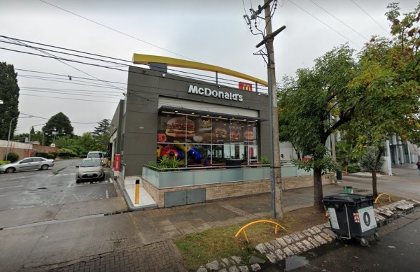McDonald's exterior con ventanas grandes, menú visible, coches estacionados y árboles alrededor. Día nublado.