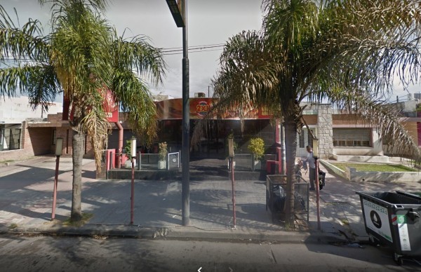 Entrada de Di Metro con palmeras, farola y basurero. Fachada roja y vidriada. Calle desierta, día nublado.