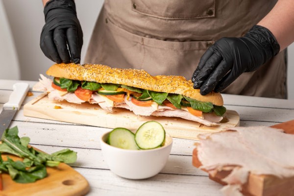 Persona con guantes negros prepara un sándwich largo con vegetales y carne sobre una mesa blanca.