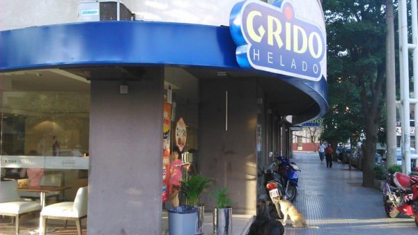 Fachada de Grido Helado con terraza, sillas blancas y motos estacionadas frente a la entrada. Árboles al fondo.