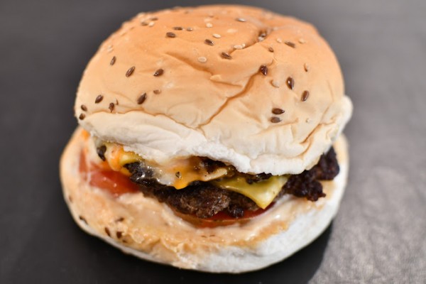 Una hamburguesa apetitosa con una cebolla crujiente y dorada en la parte superior, seguida por una rebanada de queso derretido, una jugosa carne de res, una rodaja de tomate fresco y una capa de salsa especial. Todo esto está contenido entre dos panes de 