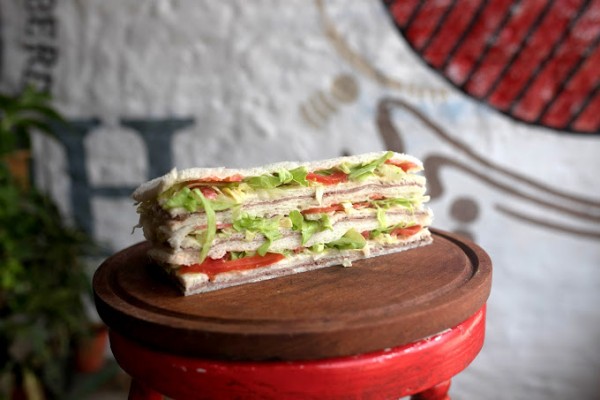 Sandwich apilado con pan blanco, lechuga y tomate, sobre taburete rojo. Fondo artístico blanco y rojo.