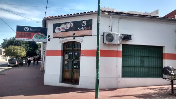 Edificio blanco con franja roja, dos carteles de sushi, puerta de entrada y persiana verde cerrada. Calle con árboles y coches.