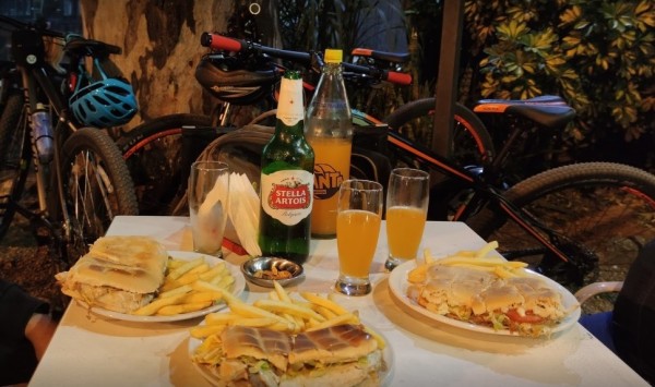 Comida en Uggo: sándwiches, papas fritas y cervezas sobre mesa blanca, bicicletas de fondo. Ambiente relajado al aire libre.