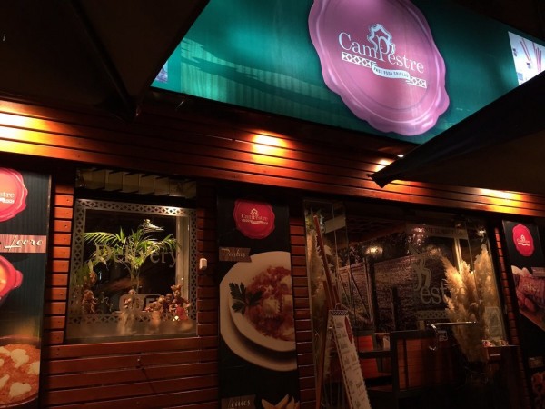 Fachada de Campestre FastFood con iluminación cálida, ventanales y cartel publicitario de comida. Ambiente acogedor y rústico.
