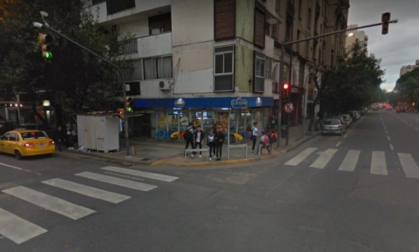 Calle con edificios, tiendas y semáforos. Gente camina cerca de Grido. Taxi amarillo y autos estacionados. Atardecer urbano.