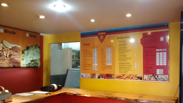 Interior de Central G con menú de pizzas, sandwiches y empanadas en pared amarilla. Iluminación brillante y mostrador de madera.