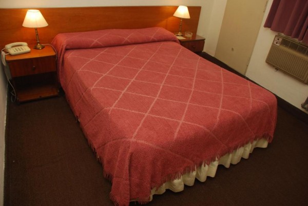 Habitación del Gran Crisol con cama grande, manta roja cuadriculada, mesitas de noche y lámparas. Ambiente acogedor.